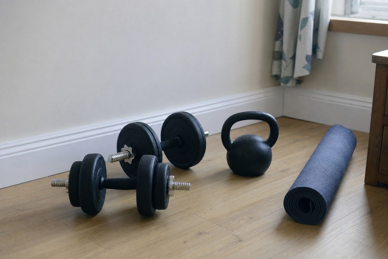 A pair of adjustable dumbbells and a kettlebell on a wooden floor in a plain home room